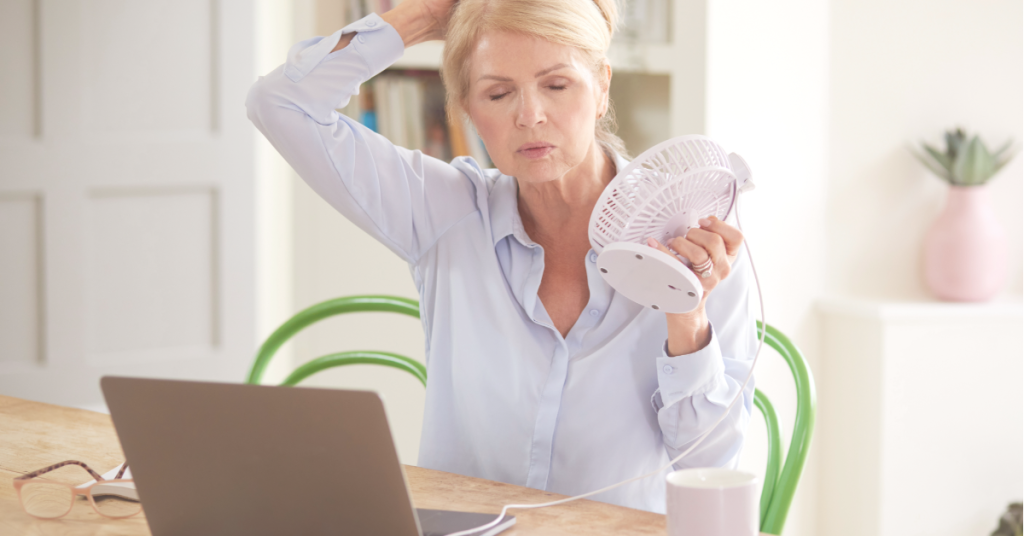 woman sitting her desk holding a mini fan to her face having hot flashes